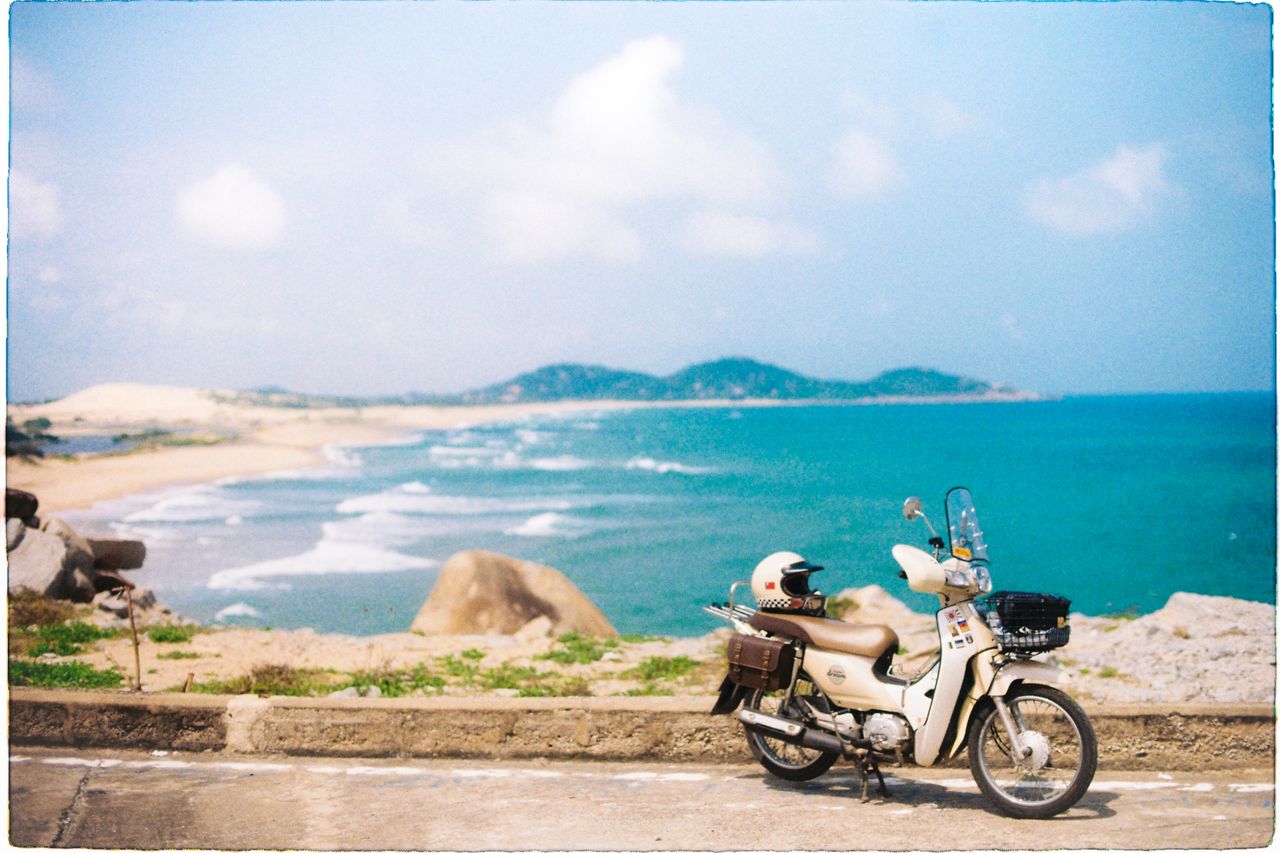 A scooter parked at a coastal roadside with a helmet resting on the seat, overlooking the sea